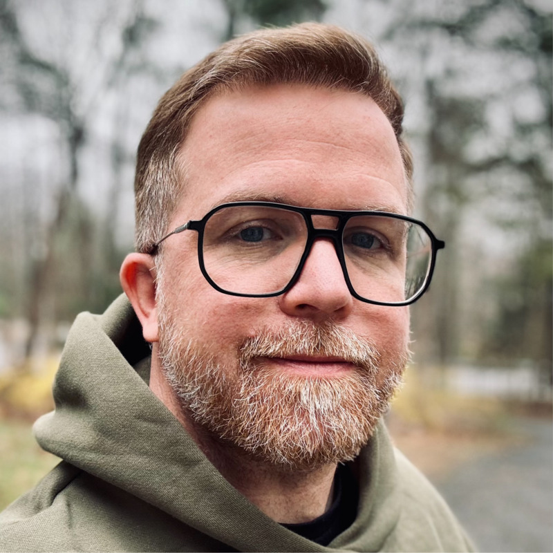 Portrait of Nathan, in a green hoodie and dark-framed aviator glasses, outdoors.